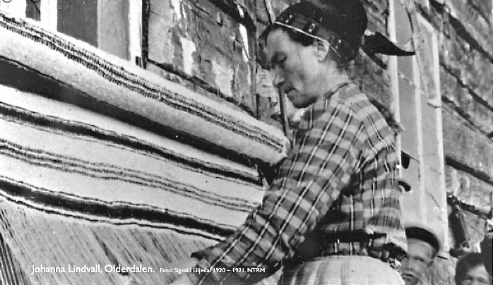Johanna Lindvall vever grene i Dálošvággi/ Olderdalen, Gáivuotna/ Kåfjord. Greneveven er satt opp mot husets yttervegg. Fotografiet er tatt ca. 1920 av Sigvald Liljedal. Nord Troms museum.